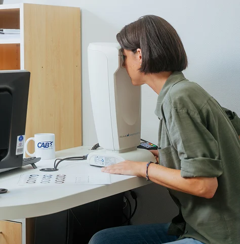 Frau schaut durch ein Sehhilfegerät an einem Schreibtisch mit Computer und Tasse.