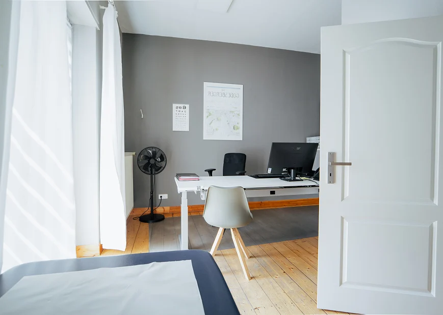 Helles Untersuchungszimmer mit Schreibtisch, Stuhl und medizinischen Utensilien für Vorsorgeuntersuchungen.