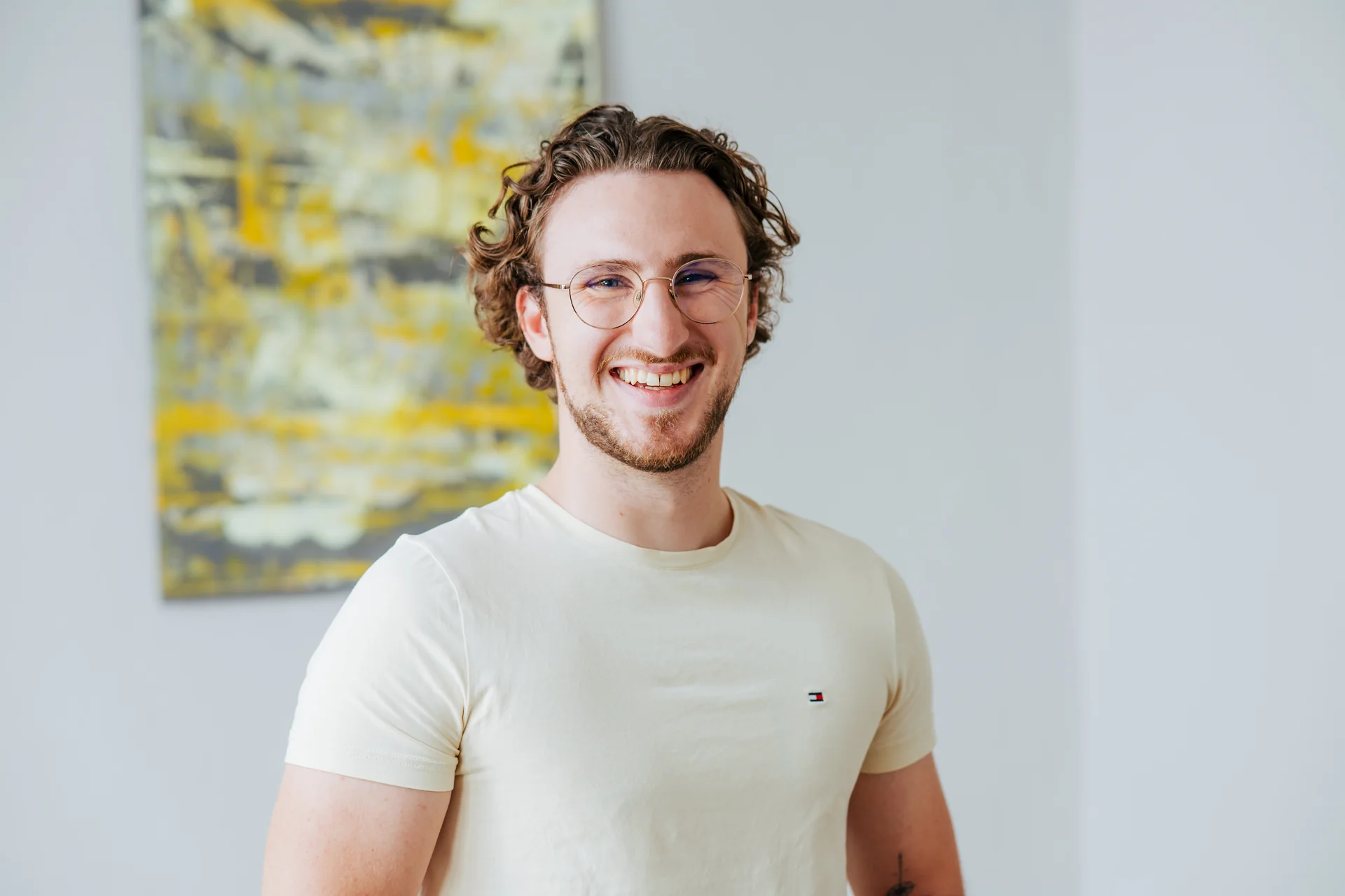 Lächelnder junger Mann mit lockigem Haar und Brille vor einem modernen Kunstwerk.