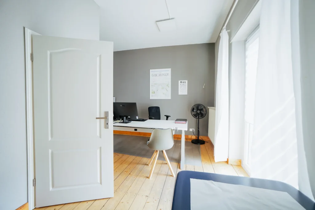 Büro mit Schreibtisch, Computer und Stuhl, sichtbar durch eine offene Tür in einem modernen Raum.