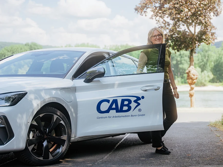 Frau steht neben einem weißen Auto mit dem Logo des Centrums für Arbeitsmedizin Bonn GmbH.
